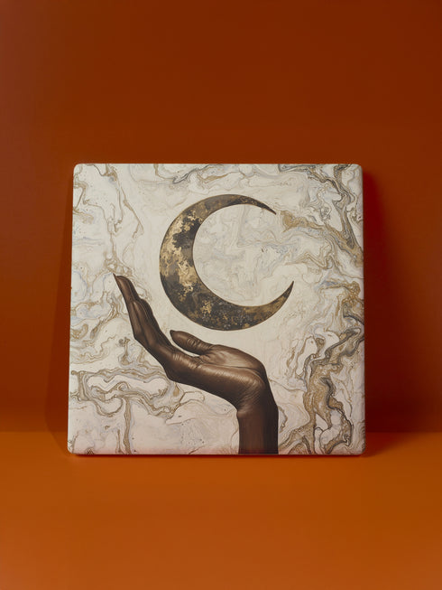 A ceramic coaster featuring a bronze-colored hand reaching toward an antiqued gold crescent moon against a swirling ivory and gold marble background, available in round and square shapes with cork backing.
